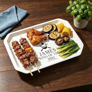 Op de afbeelding: Een wit rechthoekig serveerblad met gegrild voedsel, waaronder kip, spiesjes, maïs, aubergine en champignons. Het blad heeft de tekst "JAMES KING OF THE BBQ" in het zwart. Een blauwe servet en een potplant staan op de achtergrond.