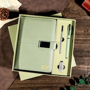 May include: A sage green journal and pen set in a gift box. The journal has a magnetic clasp and the initials "KY" in gold. The set includes a pen, a refill, and a silver-tone clip. The box is open, revealing the contents.