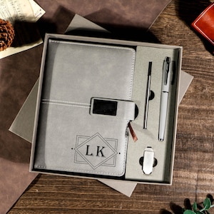 May include: A gray journal and pen set in a gift box. The journal has a geometric design with the initials "LK". The set includes a pen, a stylus, and a pen clip. The box is gray and sits on a wooden surface.