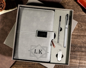 May include: A gray journal and pen set in a gift box. The journal has a geometric design with the initials "LK". The set includes a pen, a stylus, and a pen clip. The box is gray and sits on a wooden surface.