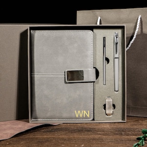 Puede incluir: Un juego de cuaderno y bolígrafo gris en una caja de regalo. El cuaderno tiene un cierre rectangular plateado y las iniciales "WN" en dorado. El juego incluye un bolígrafo, un lápiz óptico y un portalápices. La caja de regalo está en una bolsa de regalo marrón más grande.