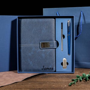Pode incluir: Um conjunto de diário e caneta de couro azul apresentado numa caixa de presente. O diário tem um fecho e o nome "James" está gravado na capa. Inclui-se uma caneta azul, uma caneta prateada e um clip para caneta prateado.