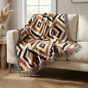 May include: A woven throw blanket featuring a geometric pattern of diamonds and zigzags in shades of cream, tan, brown, and black. The blanket has a textured surface and is finished with decorative fringe along the edges, draped over a sofa.