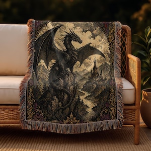 May include: A woven throw blanket featuring a detailed illustration of a black dragon perched on a rock overlooking a castle and a river. The blanket has a fringed edge and a vintage aesthetic, with a color palette of black, brown, and gold.