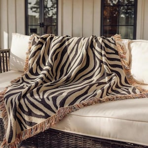 Pode incluir: Uma manta tecida com um padrão de riscas de zebra em preto e bege de alto contraste. O têxtil retangular tem uma construção de malha texturizada e é rematado com bordas decorativas de franjas multicoloridas nos quatro lados.