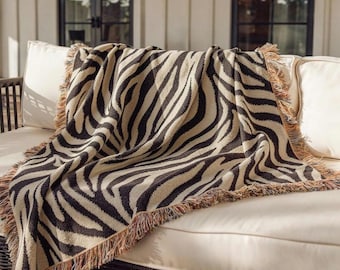 May include: A woven throw blanket featuring a high-contrast black and beige zebra stripe pattern. The rectangular textile has a textured knit construction and is finished with decorative multi-colored fringed edges along all four sides.