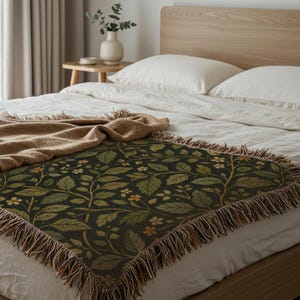 May include: A dark green throw blanket with a botanical pattern of leaves and small flowers in shades of green and gold. The blanket has a fringed edge and is draped over a bed with white linens.