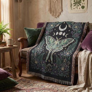 May include: Black woven throw blanket with a large green moth design, moon phases, and floral details. The blanket has a fringed edge and is draped over a chair with green and purple cushions. The design is inspired by celestial and natural themes.
