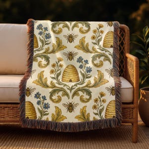 May include: A patterned throw blanket featuring bees, beehives, and floral designs in yellow, blue, and green on an off-white backdrop. The blanket has a fringed edge in brown and tan hues. It is draped over a wicker chair, suitable for home decor.