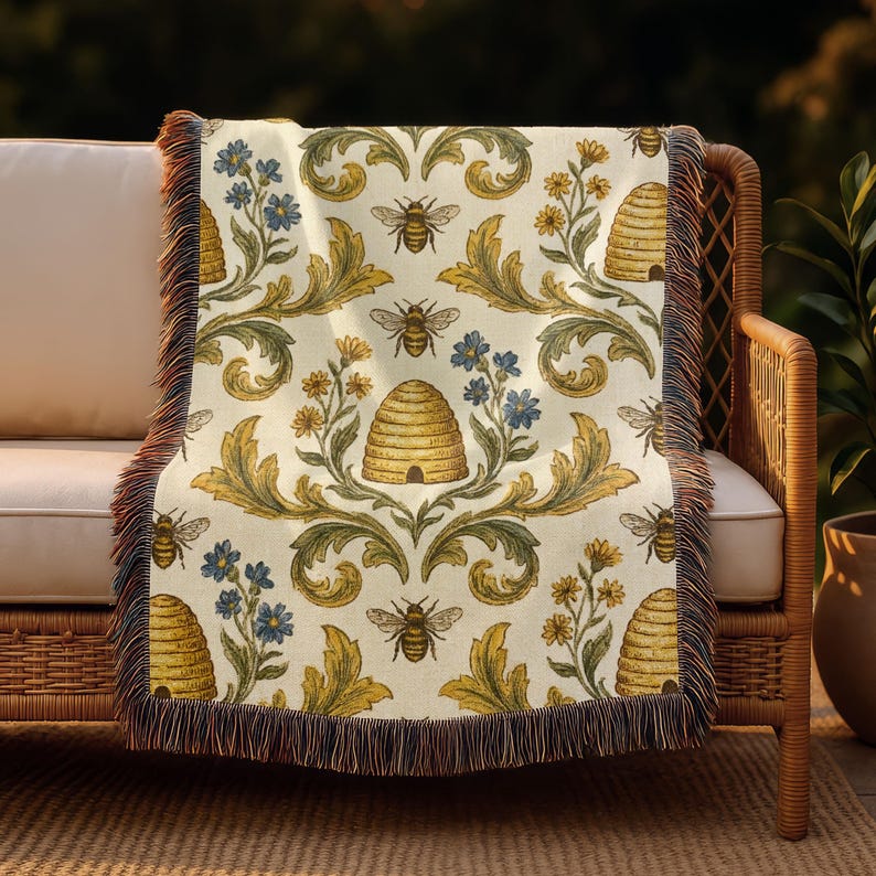 May include: A patterned throw blanket featuring bees, beehives, and floral designs in yellow, blue, and green on an off-white backdrop. The blanket has a fringed edge in brown and tan hues. It is draped over a wicker chair, suitable for home decor.