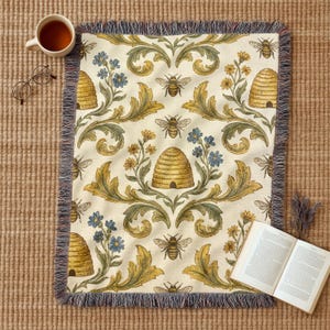May include: A cream-coloured throw blanket with a bee and beehive pattern. The blanket features a repeating design of bees, beehives, and floral elements in yellow, blue, and green. The blanket has a fringed edge and is displayed with a cup of tea, glasses, and a book.