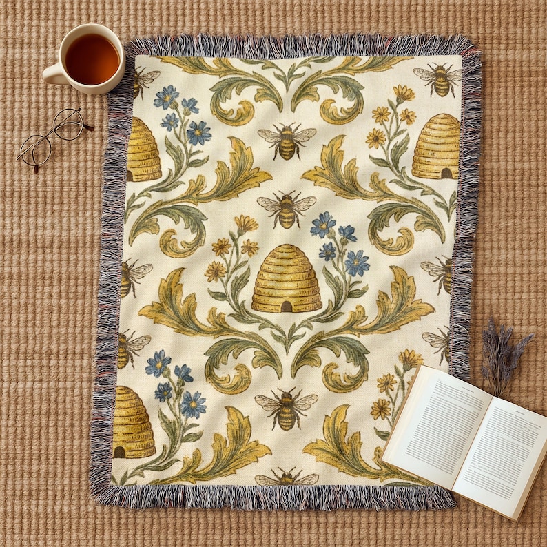 May include: A cream-coloured throw blanket with a bee and beehive pattern. The blanket features a repeating design of bees, beehives, and floral elements in yellow, blue, and green. The blanket has a fringed edge and is displayed with a cup of tea, glasses, and a book.
