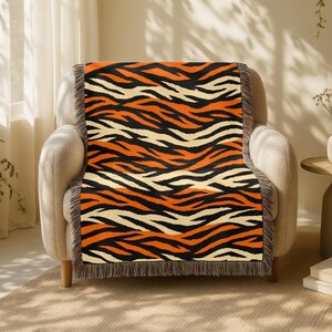 May include: A patterned throw blanket featuring a tiger stripe design in black, orange, and cream. The blanket is draped over a beige armchair. The blanket has a fringed edge.