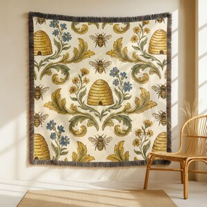 May include: A woven throw blanket featuring a repeating pattern of bees, beehives, and floral designs in yellow, blue, and green on an off-white backdrop. The blanket has a fringed edge and is displayed on a wall.