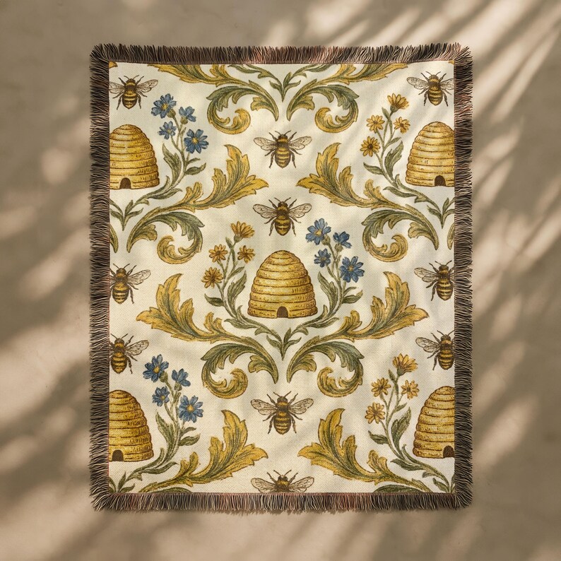 May include: Cream-coloured throw blanket featuring a repeating pattern of bees, beehives, and floral elements in yellow, blue, and green. The blanket has a fringed edge.