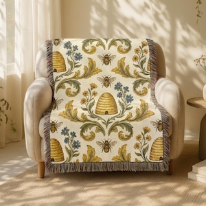 May include: A cream-coloured throw blanket draped over a beige armchair. The blanket features a repeating pattern of bees, beehives, and floral elements in yellow, blue, and green. The edges of the blanket have a fringed trim.