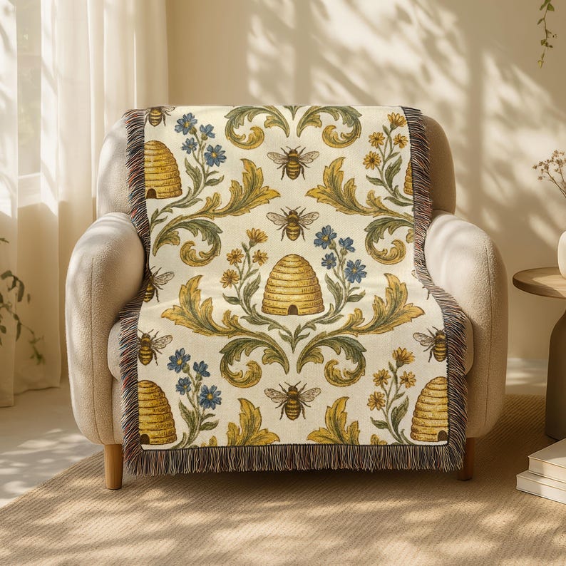May include: A cream-coloured throw blanket draped over a beige armchair. The blanket features a repeating pattern of bees, beehives, and floral elements in yellow, blue, and green. The edges of the blanket have a fringed trim.