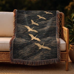 May include: A dark grey throw blanket with a gold crane design. The blanket features a repeating pattern of cranes in flight over a wavy background. The edges are finished with a fringe in shades of brown and tan.