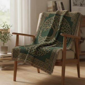 May include: A green throw blanket with a Celtic tree of life design draped over a wooden armchair. The blanket features a detailed pattern in tan and green, with a fringed edge. The chair has a light-colored cushion.