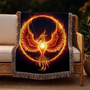 May include: A black throw blanket featuring a vibrant phoenix design. The phoenix is depicted in shades of orange and red, encircled by a fiery halo. The blanket has a fringed edge and is draped over a wicker chair.