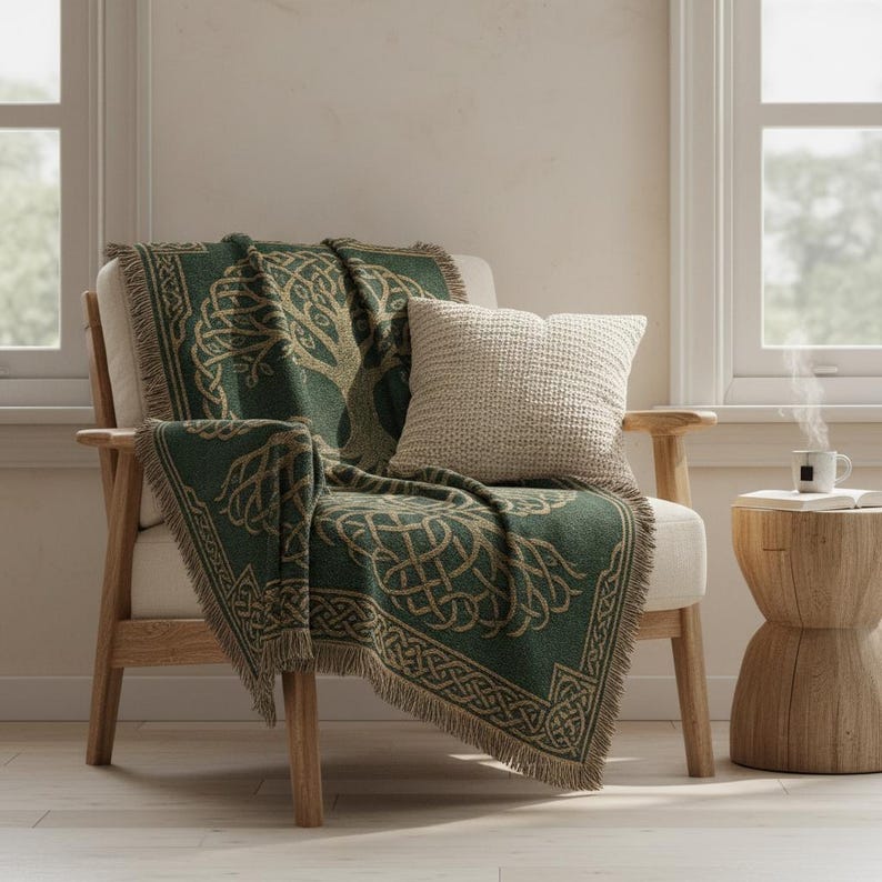 May include: A green and tan throw blanket with a Celtic tree of life design draped over a cream-colored armchair. A cream-colored square pillow rests on the chair. The blanket has a fringed edge. A small wooden side table holds a cup of tea.