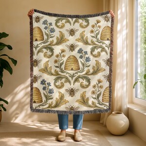 May include: Cream-coloured throw blanket featuring a repeating pattern of bees, beehives, and floral elements in gold, green, and blue. The blanket has a fringed edge.
