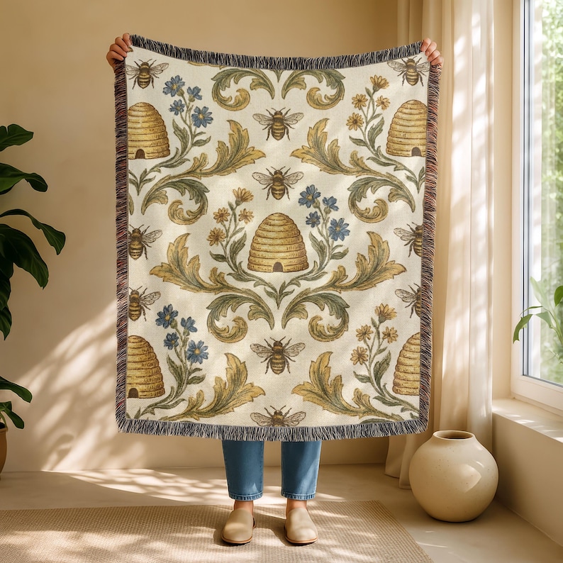 May include: Cream-coloured throw blanket featuring a repeating pattern of bees, beehives, and floral elements in gold, green, and blue. The blanket has a fringed edge.