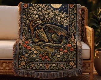 May include: A woven tapestry throw blanket featuring a green dragon resting under a flowering tree, with a full moon in the background. The blanket has a dark blue background with floral and foliage borders and fringed edges. The throw is draped over a wicker chair.