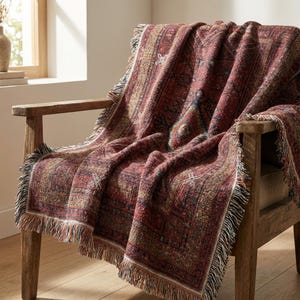 Op de afbeelding: Een rood en beige patroon plaid met franjes, gedrapeerd over een houten stoel. De deken heeft een geometrisch ontwerp met een vintage esthetiek, wat warmte en textuur aan de ruimte toevoegt.