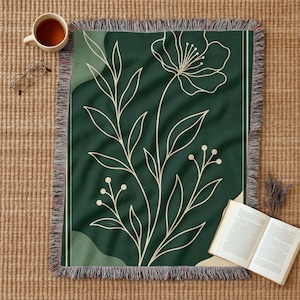 Op de afbeelding: Een donkergroene sprei met een crèmekleurig bloemmotief en franjes. De sprei heeft een botanische illustratie van een bloem en bladeren. Een kopje thee, een bril en een open boek liggen op een geweven oppervlak.