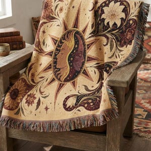 May include: A woven throw blanket featuring a central sun and moon face motif in shades of gold, cream, and deep burgundy. The design includes intricate floral patterns, stars, and decorative swirls, finished with a fringed edge on all sides.