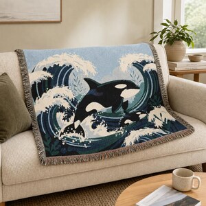 May include: A woven throw blanket featuring a large black and white orca whale leaping through stylized dark blue and white ocean waves. The rectangular blanket has a fringed edge and displays a detailed graphic design of marine life.