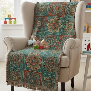May include: A teal throw blanket with a floral pattern in shades of orange, yellow, and brown is draped over a beige armchair. The blanket has a fringed edge. Wooden toy blocks and houses are on the chair.