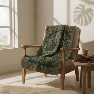 May include: A green throw blanket with a Celtic tree of life design draped over a wooden armchair. The blanket has a fringe edge and a detailed knotwork border. The chair is next to a small wooden side table with books and a mug.