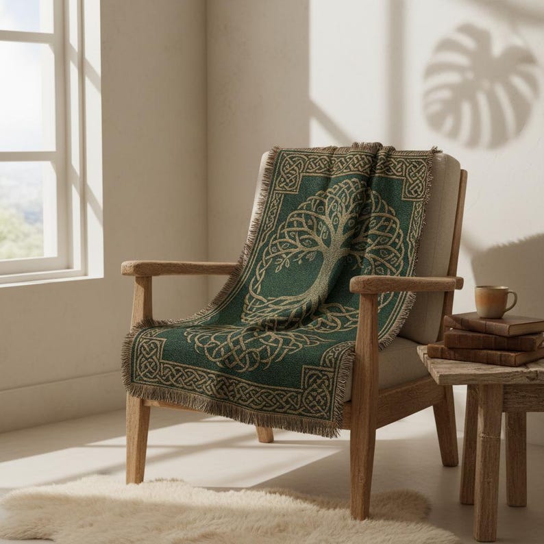 May include: A green throw blanket with a Celtic tree of life design draped over a wooden armchair. The blanket has a fringe edge and a detailed knotwork border. The chair is next to a small wooden side table with books and a mug.