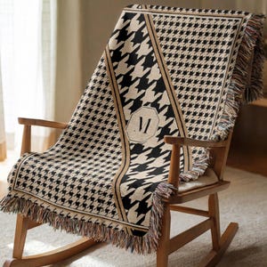 May include: A woven throw blanket featuring a black and beige houndstooth pattern with a central vertical stripe and a hexagonal emblem containing the letter M. The blanket has fringed edges and is draped over a wooden rocking chair.
