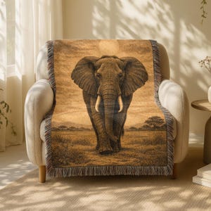 Op de afbeelding: Een geweven sprei in sepia-tinten met een illustratie van een olifant. De sprei, met een gefranjerde rand, is over een beige fauteuil gedrapeerd. De achtergrond toont een savannelandschap met bomen en horizon.