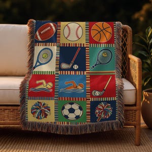 May include: A woven throw blanket featuring a patchwork design with various sports illustrations. The squares depict a football, baseball, basketball, tennis, golf, swimming, soccer, and cheerleading pom-poms. The blanket has a fringed edge and is draped over a wicker chair.