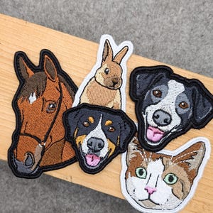 Könnte beinhalten: Gestickte Aufnäher mit einem Pferd, einem Kaninchen, zwei Hunderassen und einer Katze. Die Aufnäher haben schwarze Ränder und sind auf einem hellen Holzgrund platziert. Das Pferd ist braun, das Kaninchen ist hellbraun, und die Hunde und die Katze haben verschiedene Muster.