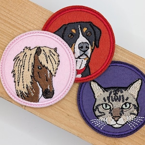 Könnte beinhalten: Drei kreisförmige gestickte Aufnäher mit Tierporträts. Ein Aufnäher ist hellrosa mit einem braunen Pferd, einer ist rot mit einem schwarz-weiß-braunen Hund und einer ist lila mit einer grau-weißen getigerten Katze mit grünen Augen.