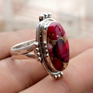 Puede incluir: Un anillo de plata con una piedra preciosa ovalada roja y dorada. La piedra preciosa está engastada en un bisel decorativo de plata con pequeños detalles de cuentas. La banda del anillo es de plata pulida.