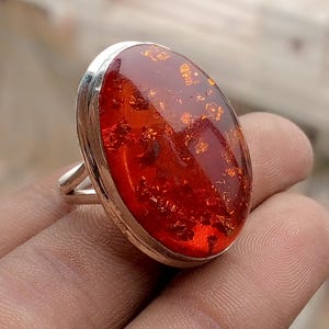 May include: Oval-shaped amber ring with a silver band. The amber stone is a deep reddish-orange color with visible inclusions. The ring is set in a polished silver bezel and has a simple, elegant design. A piece of jewelry.