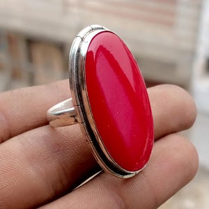 May include: A silver ring with a large, oval, bright red gemstone. The stone is set in a silver bezel with a detailed edge. The ring is held in a hand, highlighting the polished stone's colour and design.