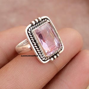 Peut inclure: Bague en argent avec une pierre précieuse rectangulaire rose clair. La pierre est sertie dans un cadre décoratif avec un motif de corde torsadée et de petits accents perlés. La bague est sur le doigt d'une personne.