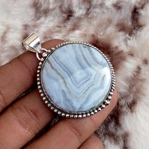 Può includere: Un ciondolo rotondo con una pietra preziosa di agata blu a pizzo lucidata con bande bianche e azzurre. La pietra è incastonata in una cornice in argento sterling con un bordo perlato e un semplice gancio in argento sulla parte superiore.