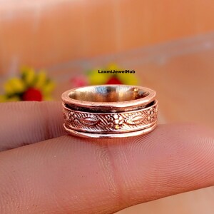 Può includere: Un anello spinner color rame caratterizzato da una fascia centrale con un motivo floreale e a foglie in rilievo. L'anello presenta due bordi esterni lisci e rialzati che incorniciano la sezione centrale testurizzata, che ruota indipendentemente.