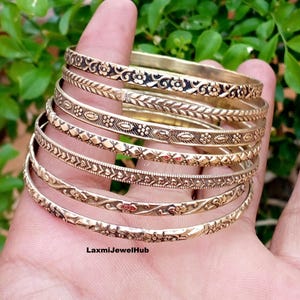 Pode incluir: Um conjunto de seis pulseiras empilhadas de metal em tom dourado com vários padrões em relevo, incluindo motivos florais, formas geométricas e texturas de folhas. Cada pulseira tem um design gravado distinto com detalhes escuros em baixo-relevo.