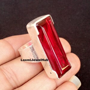May include: A silver ring featuring a large, rectangular, deep red gemstone. The gemstone is set in a silver band with a rectangular top. The ring is held in a hand against a dark background. The text "LaxmiJewelHub" is visible on the ring.