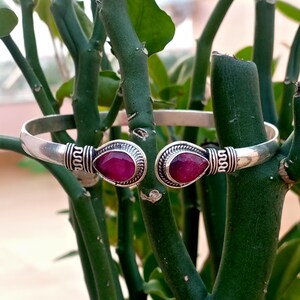 May include: A silver cuff bracelet featuring two teardrop-shaped, faceted deep pink gemstones at the open ends. The metal band displays intricate, textured silver detailing surrounding each stone, providing a decorative contrast to the smooth finish.