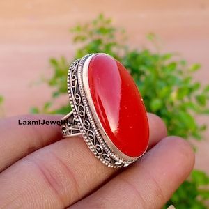 May include: A silver ring featuring a large, smooth, oval-shaped red stone set in a decorative metal frame with intricate wirework patterns along the sides. The band displays a detailed filigree design, and the text LaxmiJewelHub is visible.